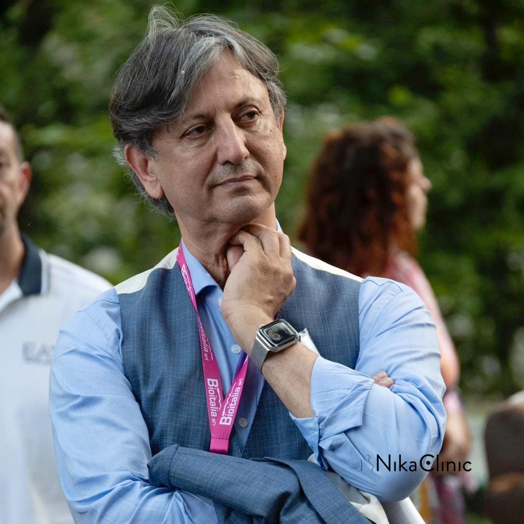 Un uomo con i capelli grigi, una camicia e un gilet blu, un orologio e un cordino rosa, è in piedi all'aperto a Milano, con una mano sul mento e un'aria contemplativa. Dott. Jafar Hassibi della Nika Clinic di Milano.