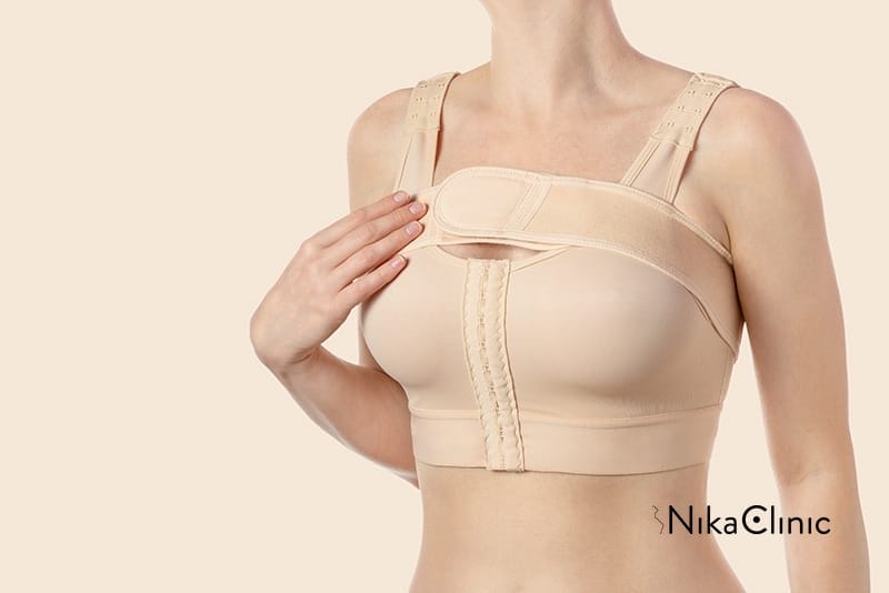 Una persona che indossa un reggiseno compressivo post-operatorio beige con spalline regolabili, toccando la cinghia anteriore con una mano, è nella fase di recupero dopo mastoplastica additiva.